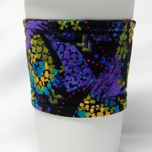 May include: A white paper coffee cup with a lid, wrapped with a fabric sleeve. The sleeve is black with a colorful pattern of purple, yellow, and teal splotches.