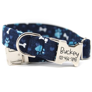 May include: A navy blue dog collar with a pattern of white bones and blue paw prints. The collar has a silver metal buckle and a name tag that reads "Buckey" and a phone number.