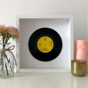 May include: A framed black vinyl record with a yellow label. The label has the text "ABBA" and "Ring Ring" on it. The record is in a white frame with a white mat.