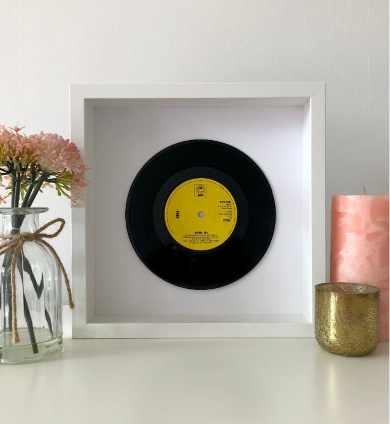 May include: A framed black vinyl record with a yellow label. The label has the text "ABBA" and "Ring Ring" on it. The record is in a white frame with a white mat.