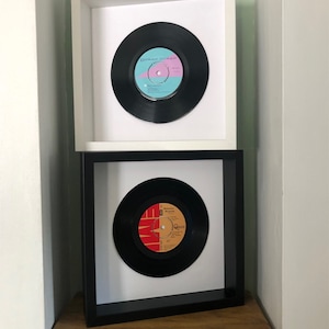 May include: Two framed vinyl records on a wooden shelf. The top frame is white with a black vinyl record featuring the text "Duran Duran". The bottom frame is black with a white vinyl record featuring the text "Queen".