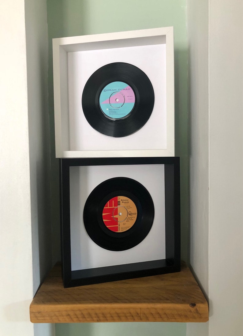 May include: Two framed vinyl records on a wooden shelf. The top frame is white with a black vinyl record featuring the text "Duran Duran". The bottom frame is black with a white vinyl record featuring the text "Queen".