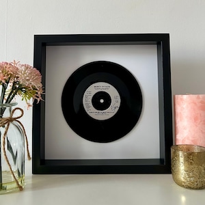 May include: A framed black vinyl record, featuring the text "Monty Python" and "Always Look on the Bright Side of Life." The record is set against a white background within a black square frame. There is a vase of pink flowers and a pink candle.