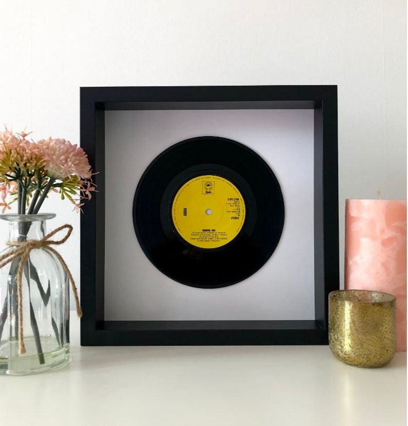 May include: A framed black vinyl record with a yellow label. The label has the text "ABBA" and "Knowing Me, Knowing You" on it. The record is in a black frame with a white mat.