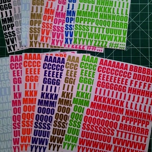 May include: An assortment of colourful alphabet stickers in various sizes. The stickers are arranged in rows and columns on a green cutting mat. The letters are in uppercase and include the letters A through Z, as well as numbers and symbols.