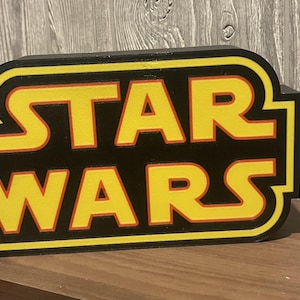 May include: A black and yellow Star Wars logo sign. The sign features the words "STAR WARS" in yellow letters with an orange outline, set against a black background. The sign is rectangular with rounded corners.