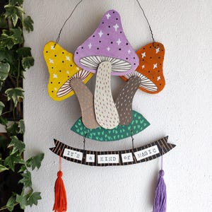 Può includere: Una decorazione da parete sospesa con tre funghi in legno dipinti in giallo, viola e arancione con dettagli a stella bianchi. Sotto i funghi, uno stendardo nero con il testo bianco IT'S A KIND OF MAGIC, rifinito con due nappe pendenti.