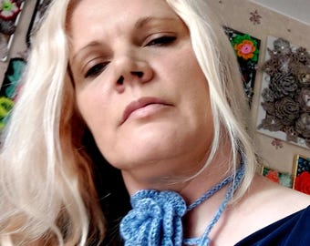 Può includere: Collana girocollo a fiore all'uncinetto, blu chiaro. Il fiore è centrato su un cordoncino intrecciato che si annoda sulla schiena. La collana è indossata con un top blu scuro.