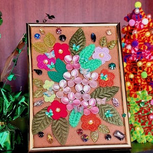 Può includere: Un'opera d'arte incorniciata che presenta un design floreale realizzato con fiori e foglie di tessuto. I fiori sono rosa, rossi e arancioni, e le foglie sono verdi. L'opera d'arte è su uno sfondo marrone e include piccole decorazioni colorate.