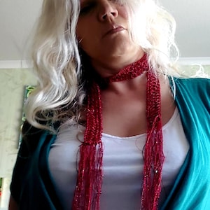May include: A long, thin, red crocheted scarf featuring a metallic, shimmering texture. The scarf is wrapped around the neck and hangs down in two long, fringed ends. The material appears to be a fine, glittery yarn with a loose, open-stitch pattern.