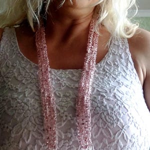 May include: A long, light pink crocheted scarf featuring integrated shimmering sequins throughout the knit. The scarf has a loose, open-weave texture and terminates in long, thin, matching pink fringe strands at both ends of the accessory.
