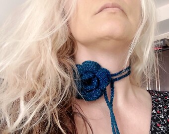 Può includere: Una collana girocollo blu all'uncinetto con un grande motivo floreale. La collana ha una chiusura con fiocco ed è adornata con perline colorate. Il fiore e la collana hanno un aspetto scintillante.