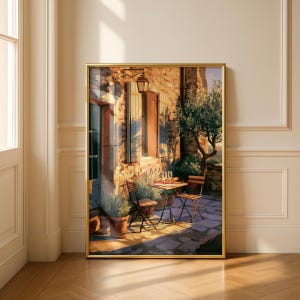 Op de afbeelding: Een ingelijste kunstdruk met de buitenkant van een zonnig stenen gebouw met een klein tafeltje en twee stoelen. De scène bevat potplanten, een lantaarn en twee wijnglazen op tafel, uitgevoerd in warme goud- en bruintinten.