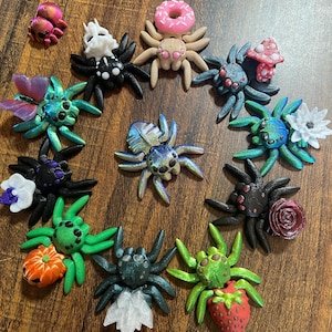 May include: A collection of 15 handmade polymer clay spider charms, each with a unique design and color scheme. The spiders feature various embellishments, including flowers, donuts, mushrooms, and pumpkins.
