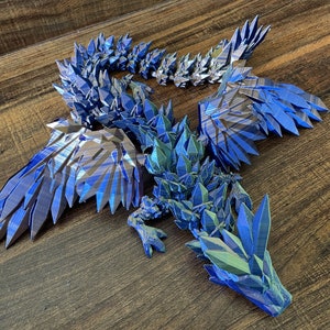 May include: A 3D printed articulated dragon figure featuring a geometric, crystal-like texture throughout its body, wings, and tail. The dragon is colored in a metallic, iridescent blue and purple gradient with sharp, angular edges and segments.