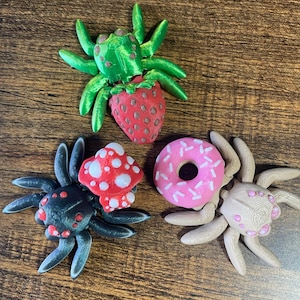May include: Three 3D-printed spider figurines. One is green with a red strawberry body, one is black with red eyes and a red-and-white mushroom on its back, and one is beige with pink eyes and a pink donut with white sprinkles on its back.