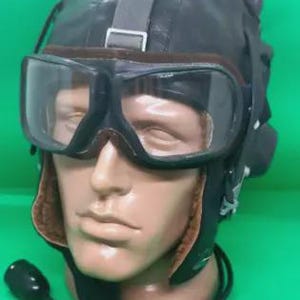 Könnte beinhalten: Ein Vintage-Fliegerhelm aus schwarzem Leder mit angebrachter Brille und Mikrofon. Der Helm hat eine braune Innenseite und einen Riemen über der Oberseite. Die Brille hat klare Gläser und einen schwarzen Rahmen. Das Mikrofon ist schwarz und mit dem Helm verbunden.
