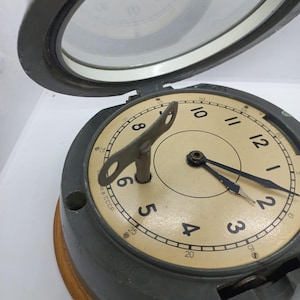 May include: An antique, round clock with a cream-colored face, black numerals, and black hands. The clock has a metal casing and a wooden base. A key is inserted in the center. The clock face is protected by a glass cover.