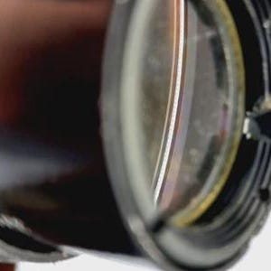 May include: Close-up of a camera lens, featuring a dark, cylindrical body with a reflective, multi-layered glass element. The lens has a black, ridged outer ring and a small, triangular opening.
