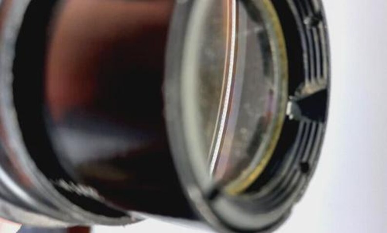 May include: Close-up of a camera lens, featuring a dark, cylindrical body with a reflective, multi-layered glass element. The lens has a black, ridged outer ring and a small, triangular opening.