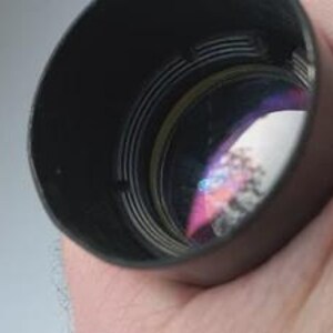 May include: A black lens hood with internal ridges, held in a hand. The lens reflects a colorful, distorted image. The hood is cylindrical and appears to be made of metal or a similar material. The background is a light, neutral color.
