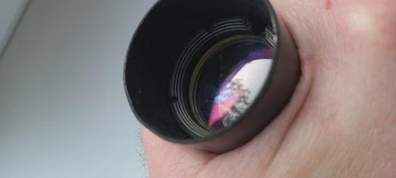 May include: A black lens hood with internal ridges, held in a hand. The lens reflects a colorful, distorted image. The hood is cylindrical and appears to be made of metal or a similar material. The background is a light, neutral color.