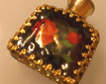 Op de afbeelding: Een kleine, vintage parfumfles met een goudkleurige metalen dop en filigraan afwerking. De fles heeft een donkere achtergrond met een geschilderd bloemmotief met rode en oranje rozen. De fles is vierkant.