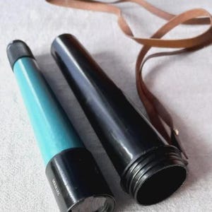 Puede incluir: Un telescopio vintage con un cuerpo negro y verde azulado, y una correa de cuero marrón. El telescopio está en dos partes, con una sección verde azulado y la otra negra. La correa de cuero está enrollada junto al telescopio.