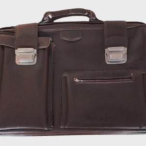 May include: A brown leather briefcase with a top handle and silver-colored clasps. The briefcase has two front pockets, one with a flap and the other with a zipper. The bag is rectangular and appears to be made of a durable material.