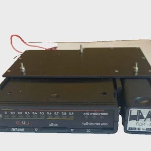 May include: A black radiation detector with a rectangular display featuring numerical readouts and a series of indicator lights. The device has a black casing and a separate rectangular component attached to the side. The text "БДРГ-35П" is visible.