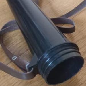 May include: A black, cylindrical object with a brown strap. The object has a wide, open base and a narrower top. The strap appears to be made of leather or a similar material. The object is lying on a wooden surface.