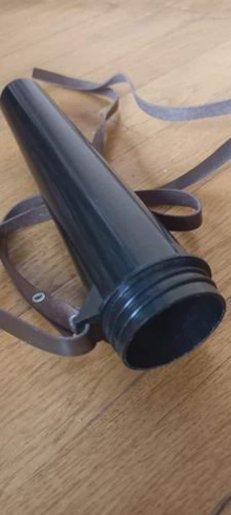 May include: A black, cylindrical object with a brown strap. The object has a wide, open base and a narrower top. The strap appears to be made of leather or a similar material. The object is lying on a wooden surface.