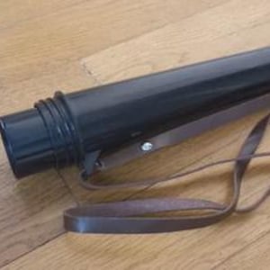 May include: A black cylindrical art portfolio case with a brown leather strap. The case is made of a hard plastic material and has a screw-on cap. The strap is attached to the case with a metal rivet.