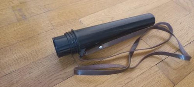 May include: A black cylindrical art portfolio case with a brown leather strap. The case is made of a hard plastic material and has a screw-on cap. The strap is attached to the case with a metal rivet.