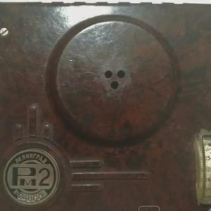 May include: A vintage brown radio receiver with a circular speaker area and a tuning dial. The face of the radio has a circular emblem with the text "ЛЕНИНГРАД" and "P2". The radio has two knobs and a small dial.
