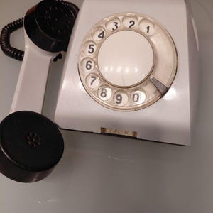 Puede incluir: Un teléfono de disco rotatorio blanco vintage con un auricular y un dial negros. El dial presenta números claros y un orificio para el dedo. El teléfono tiene una base rectangular y una etiqueta con texto.