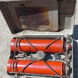 May include: Vintage camping stove with a silver metal case and two red fuel tanks. The stove has a metal frame and a round burner. The case is open, revealing the interior components. The stove is designed for outdoor cooking.