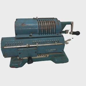 May include: Vintage teal-colored mechanical calculator with multiple number dials and a crank handle. The calculator has visible wear and tear, suggesting age and use. The text "Феликс" is visible on the top.