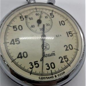 May include: A vintage mechanical stopwatch with a chrome-colored metal casing and a cream-colored dial. The face features black numerals, a sub-dial, and the Cyrillic text "Агат" and "СДЕЛАНО В СССР". Two metal push buttons are located at the top.