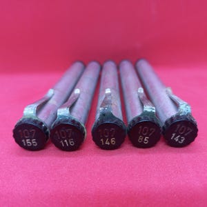 Könnte beinhalten: Nahaufnahme von fünf Metallwerkzeugen mit schwarzen Kappen und silbernen Griffen. Jede Kappe trägt die Nummer "107" und eine andere Nummer darunter. Die Werkzeuge sind vor rotem Hintergrund angeordnet.