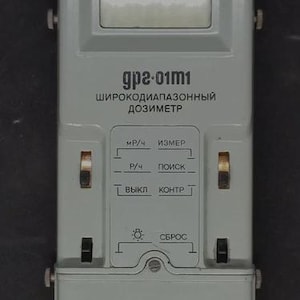 May include: A gray, rectangular dosimeter with the text "gp2-01m1" and Cyrillic text. It has a digital display, several switches, and the word "SATAPES" at the bottom. The device appears to be a radiation detector.