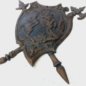 Op de afbeelding: Een bronzen decoratief schild met een reliëf van een paard en bloemmotieven. Het schild wordt gekruist door een speer en een bijl. Het ontwerp heeft een antieke, middeleeuwse uitstraling.