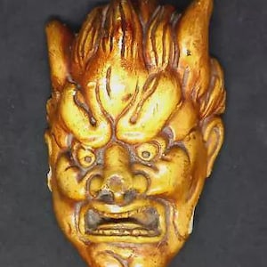 May include: A carved, golden-brown mask showing a fierce face with a wide mouth, prominent nose, and detailed hair. The mask has a textured surface, possibly made of wood. The design suggests a cultural or historical artifact, possibly from Asia.