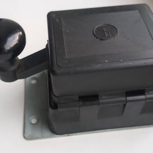 May include: A black mechanical device featuring a rounded handle and a rectangular body. The device is mounted on a metal plate with screw holes. The handle is on the left side of the device.