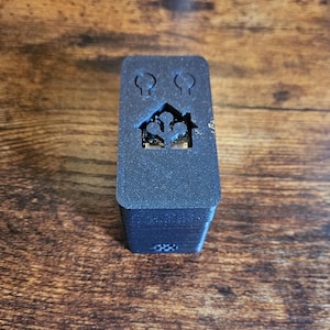 May include: A dark grey rectangular electronic device with a house and two light bulb cutouts. The device is stacked and sits on a wooden surface. The house cutout reveals a circuit board.