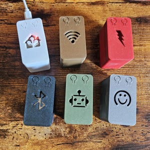May include: Six small, rectangular, white, brown, red, black, green, and gray boxes with different symbols on them. The symbols include a house, wifi symbol, lightning bolt, bluetooth symbol, robot, and smiley face. Each box has three small holes on the top.