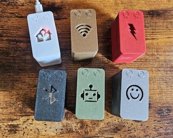 Peut inclure: Six petites boîtes rectangulaires de couleur blanche, marron, rouge, noire, verte et grise avec différents symboles. Les symboles comprennent une maison, un symbole Wi-Fi, un éclair, un symbole Bluetooth, un robot et un smiley. Chaque boîte a trois petits trous sur le dessus.