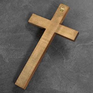 May include: A wooden cross with a natural finish, featuring a brass hanger on the back. The cross is made of light brown wood and is positioned diagonally against a dark gray background.