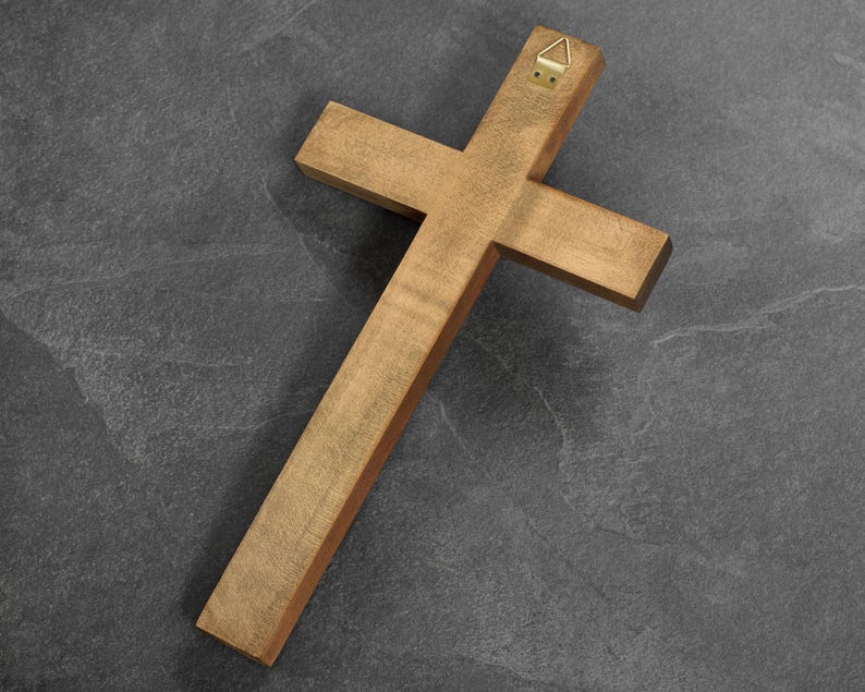 May include: A wooden cross with a natural finish, featuring a brass hanger on the back. The cross is made of light brown wood and is positioned diagonally against a dark gray background.