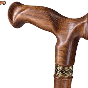 Può includere: Un bastone da passeggio in legno marrone con un'impugnatura ergonomica sagomata e una venatura del legno visibile. Sotto l'impugnatura si trova un collare metallico decorativo con un motivo a nodo celtico e la scritta ASTEROM e HANDMADE.